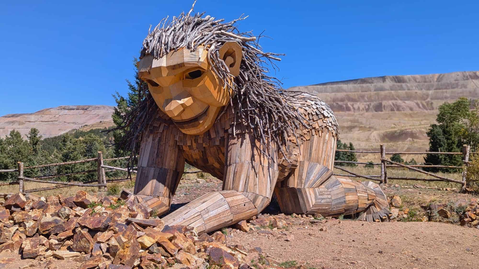 An enormous wooden troll sculpture with twig hair and a gentle expression crouches near a pile of rocks, set in a mountainous outdoor park.
