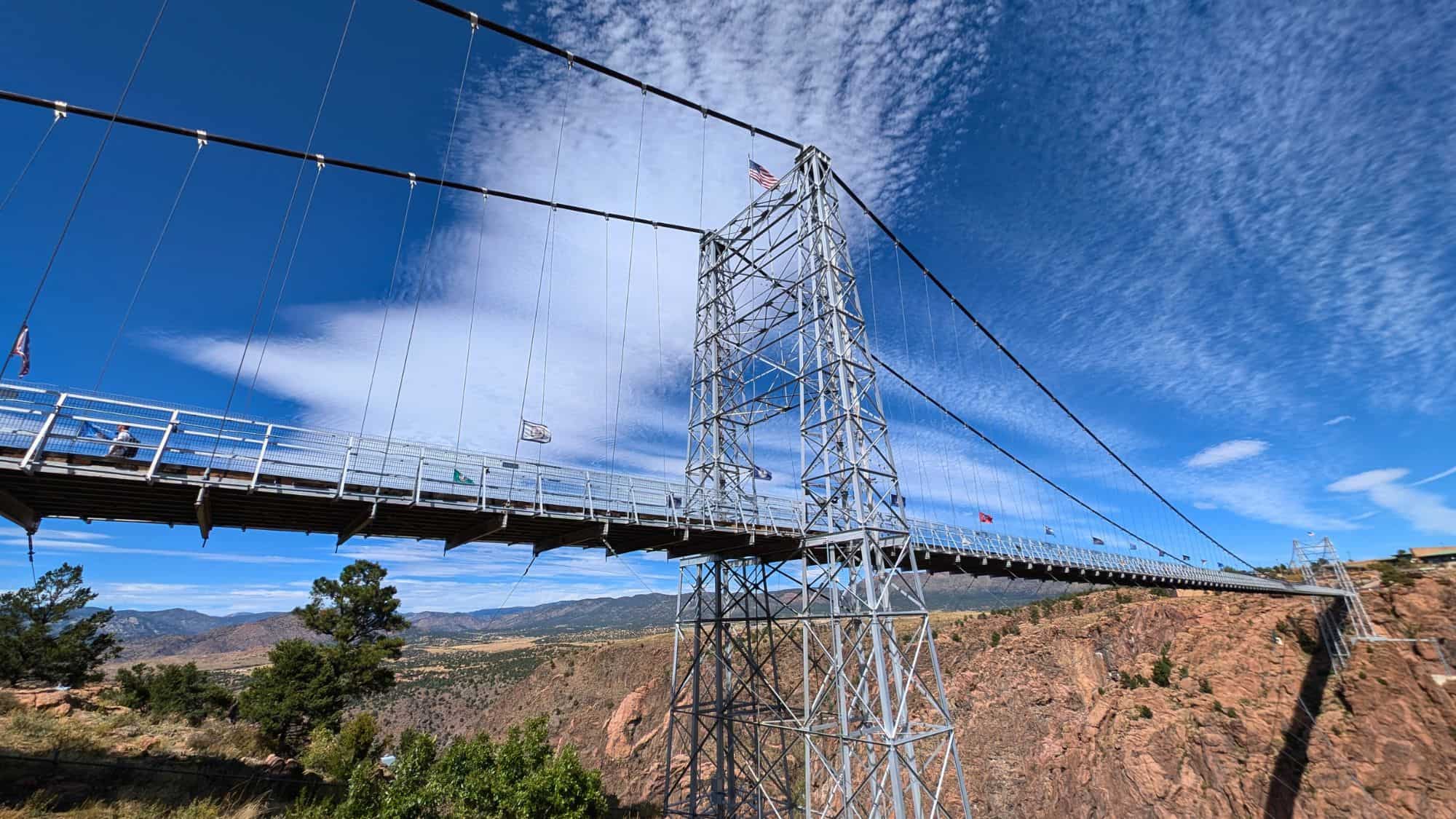 A towering suspension bridge spans a deep canyon with a metal framework, flying flags, and scenic mountain views in the distance under dramatic skies.