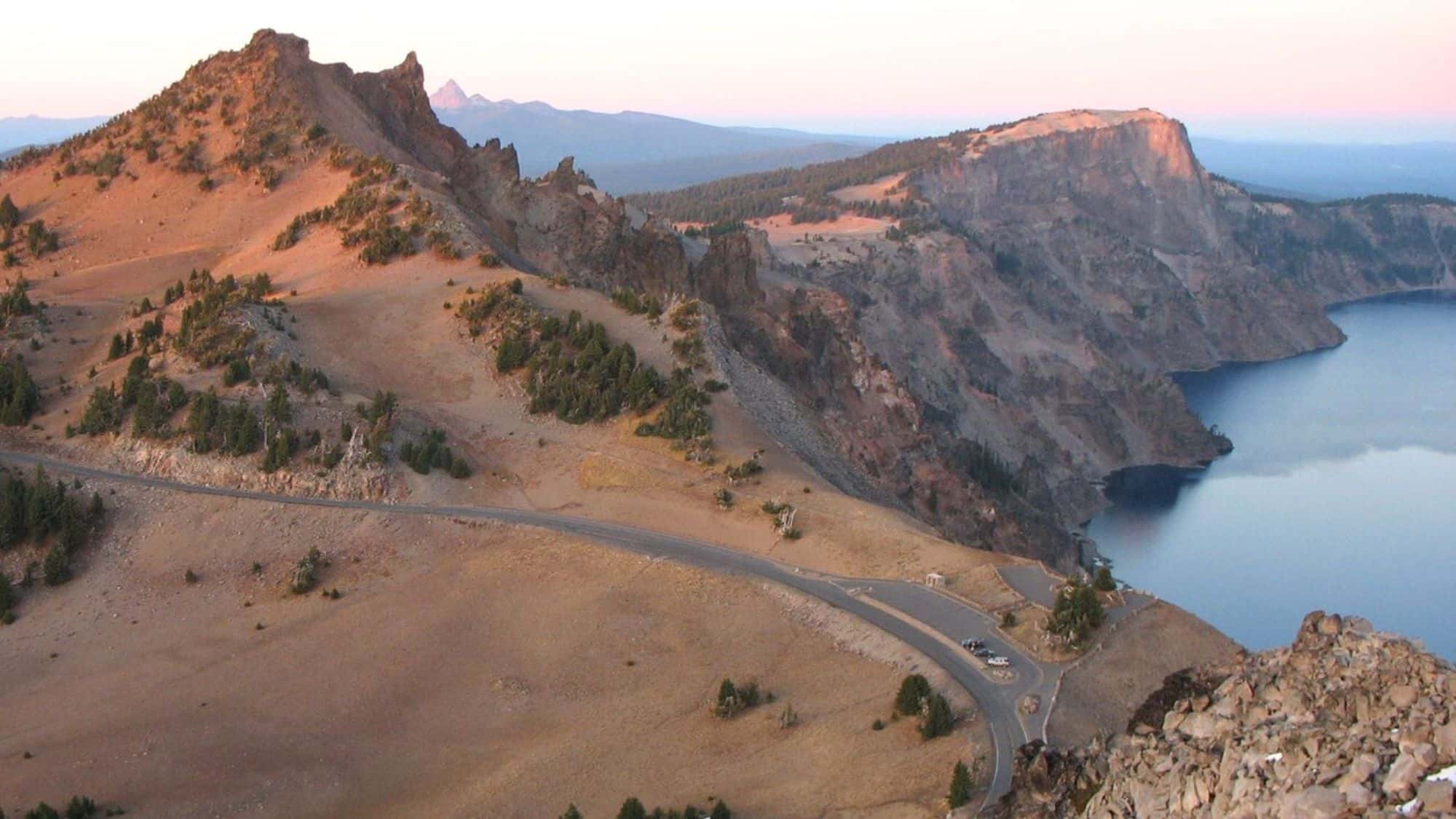 A high-elevation road curving around a rugged landscape at sunset. It offers a breathtaking view of a deep blue crater lake below, surrounded by rocky cliffs.