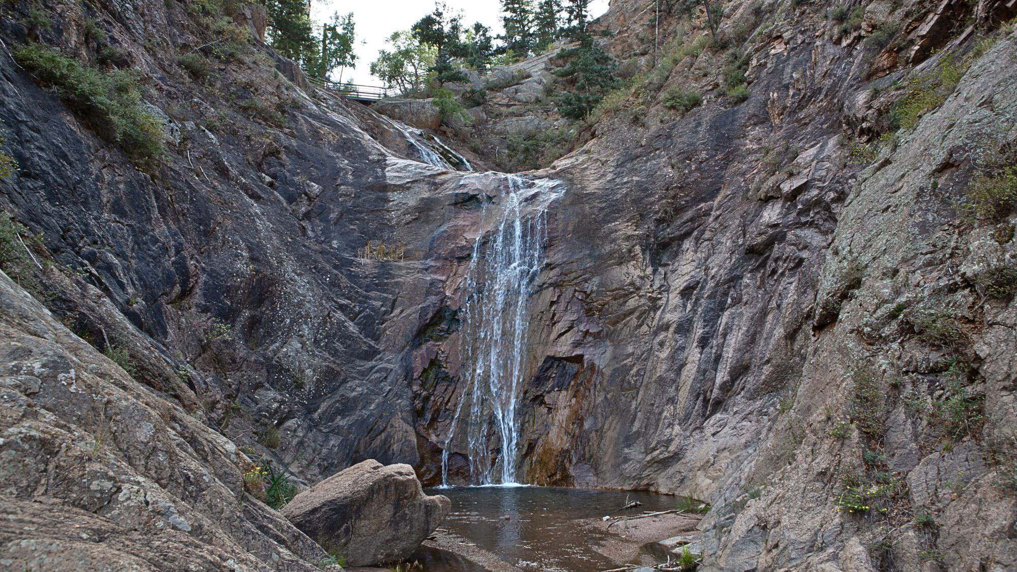 A narrow waterfall cascades down a rugged cliff face into a shallow pool surrounded by towering rock walls and sparse trees.