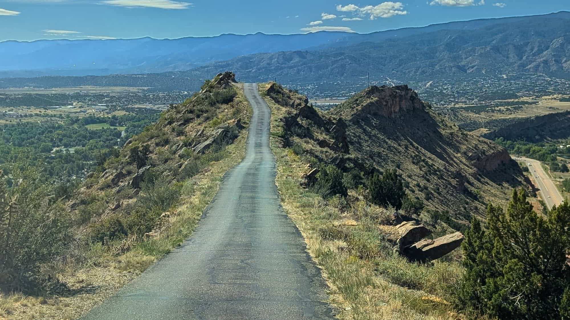 A narrow, winding road follows the ridge of a mountain with steep drop-offs on both sides, offering panoramic views of the valley and mountains below.
