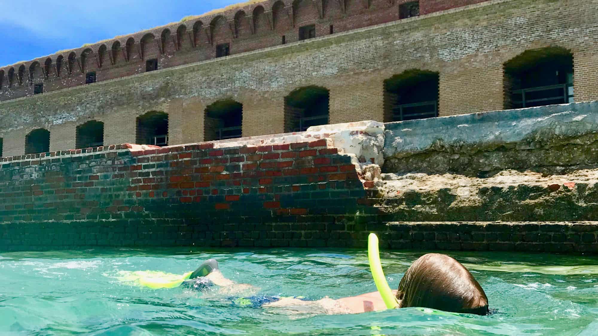 A person snorkeling in clear turquoise waters near the ruins of an old brick fort. The partially submerged red brick walls give the scene a historic charm.
