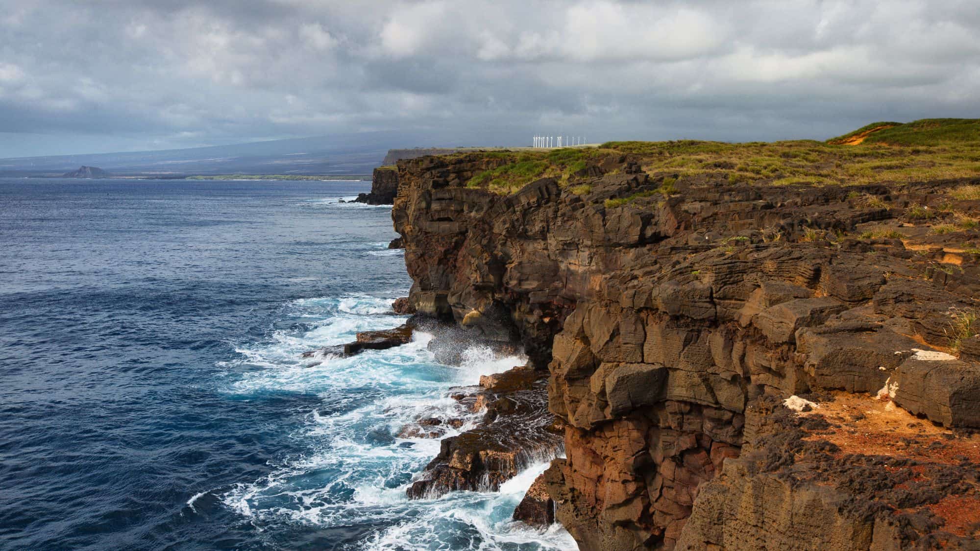 A rugged volcanic coastline where waves crash against dark cliffs. The vast ocean stretches toward the horizon, while wind turbines stand in the background under a cloudy sky.