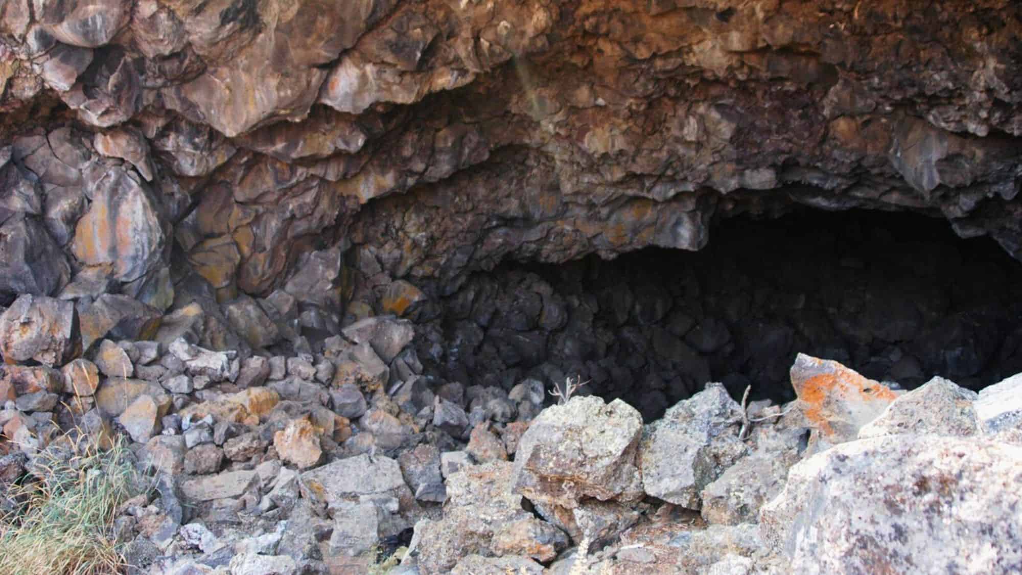 The entrance to a dark lava tube cave, surrounded by jagged volcanic rock. Loose boulders and debris cover the ground, giving the site a raw, untouched look.
