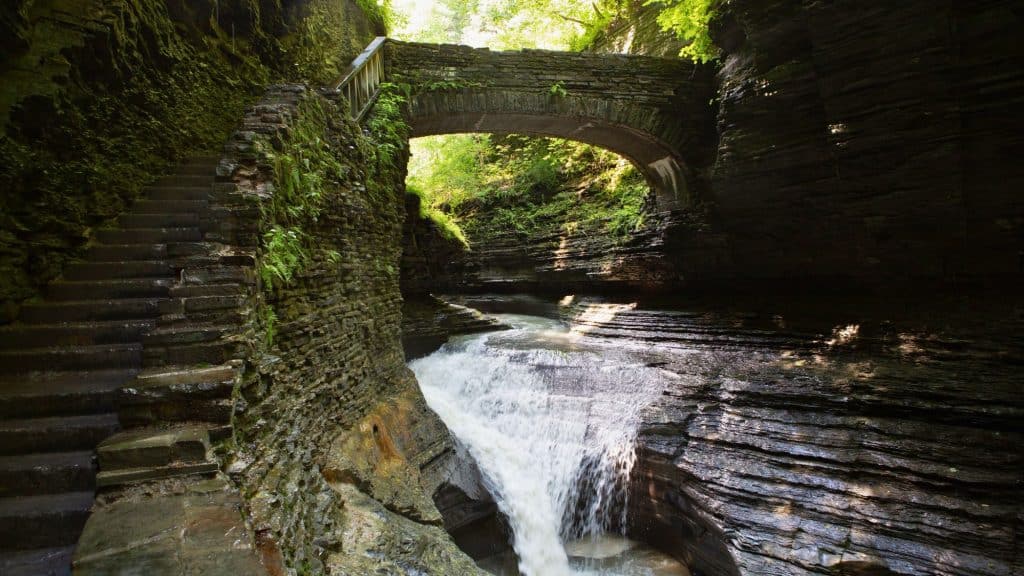 A stone bridge arches over a cascading waterfall in a lush gorge, surrounded by moss-covered rock walls and vibrant greenery. A stone staircase on the left winds up the cliffside, adding a mystical, ancient feel to the scene.