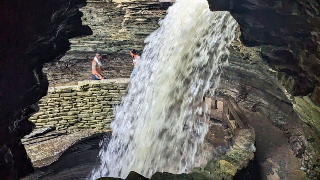 A powerful waterfall rushes down a rock face, viewed from behind through a cavernous opening. A person stands nearby on a stone ledge, dwarfed by the cascading water, creating a breathtaking perspective of the park's natural beauty.