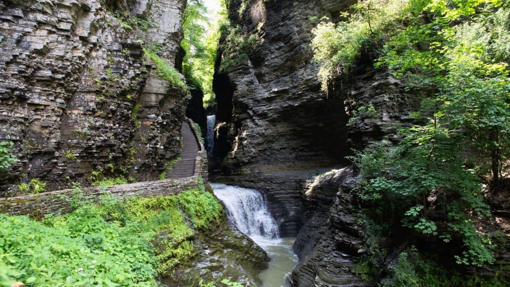 A scenic trail winds through a gorge with layered rock walls, leading to a cascading waterfall. Moss and green foliage cling to the rocks, creating a serene atmosphere.