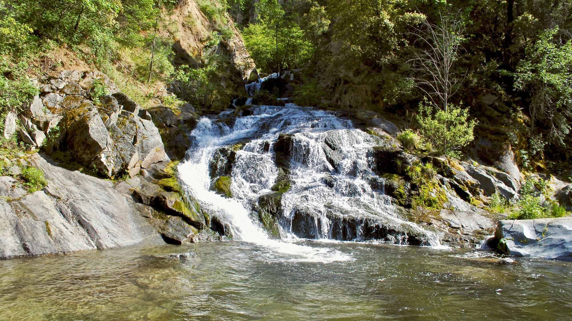 A cascading waterfall flowing over moss-covered rocks into a clear pool. The lush greenery and dappled sunlight create a refreshing, inviting scene.