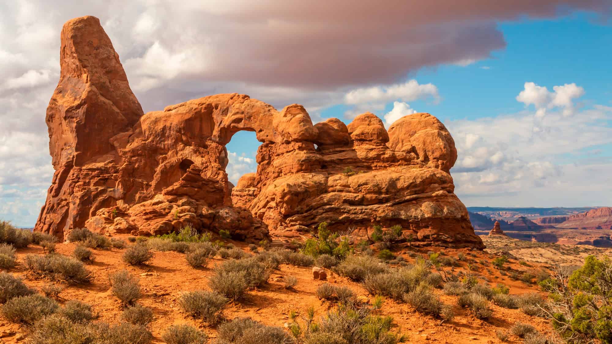 A stunning desert landscape showcasing uniquely shaped sandstone rock formations, with a prominent natural arch in the center. The sky above is a mix of blue and white, with wispy clouds stretching across the horizon.