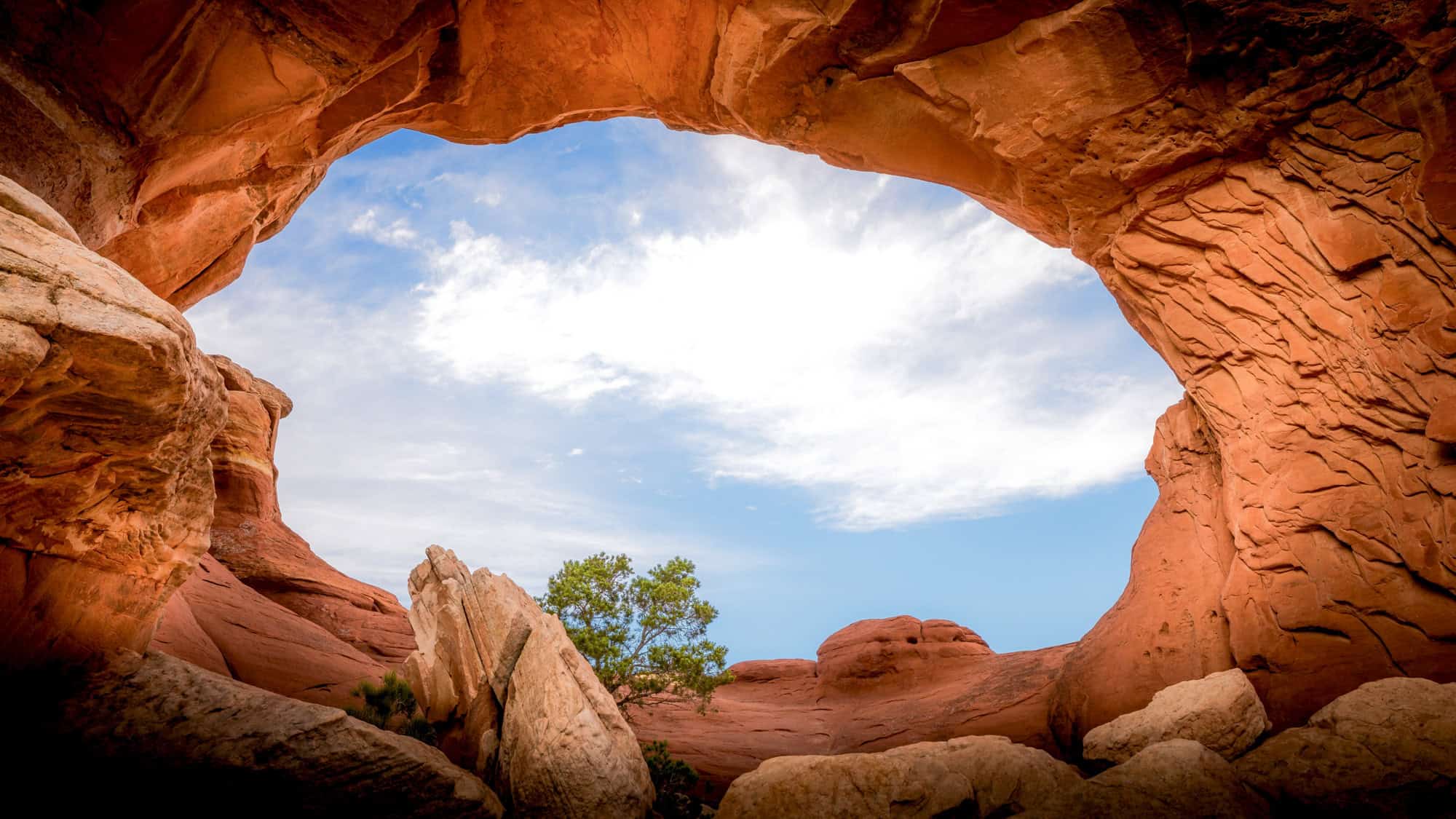 An up-close view of Broken Arch, looking through its weathered sandstone opening toward the bright sky. The rugged edges of the arch contrast with the smooth desert floor, while patches of vegetation dot the landscape.