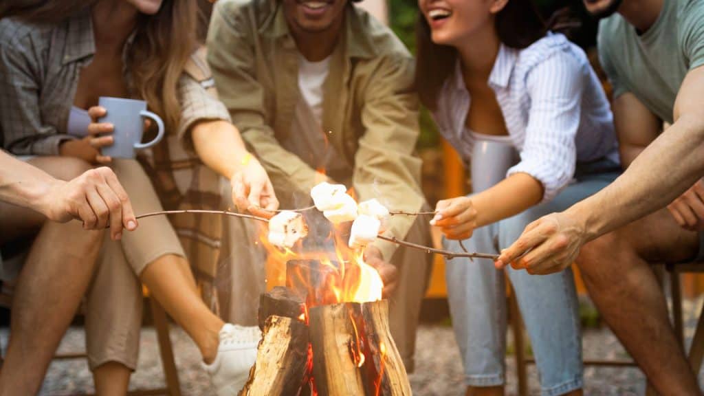 A group of friends sits around a crackling campfire, toasting marshmallows on sticks and laughing together. One person holds a warm drink, while others lean in, enjoying the cozy atmosphere of an outdoor evening.