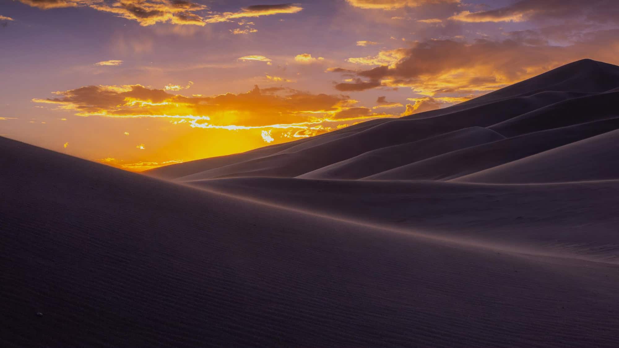 Golden sand dunes ripple under the glow of a dramatic sunset. The deep shadows and warm hues of the sky create a mesmerizing contrast in this vast and remote landscape.