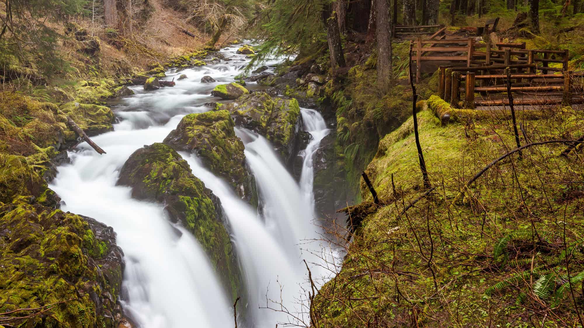 A breathtaking view of Sol Duc Falls, where multiple streams of water cascade down moss-covered rocks in a misty, forested setting. A wooden viewing platform is nestled among the trees on the right.