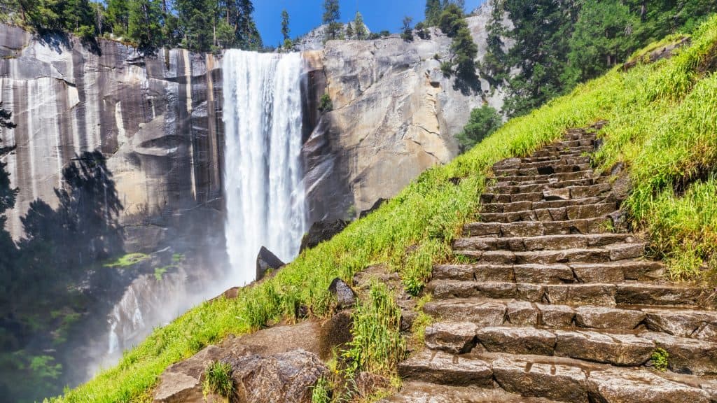 A powerful waterfall cascades down a sheer rock face, with a rugged stone staircase winding up a green hillside in the foreground. The mist from the falls creates a dramatic atmosphere against the towering cliffs.