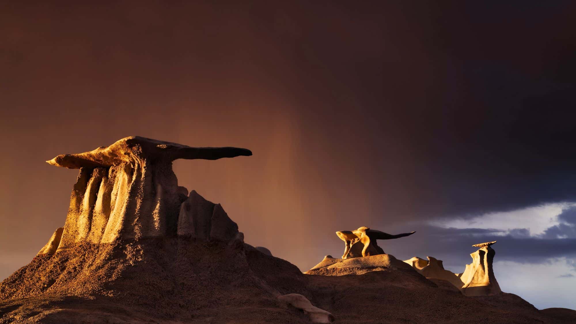 Eerie rock formations rise like petrified mushrooms in the Bisti Badlands at dusk, lit dramatically by golden hour light under a stormy sky. The dramatic contrast between the glowing rocks and dark clouds gives the scene an otherworldly vibe.