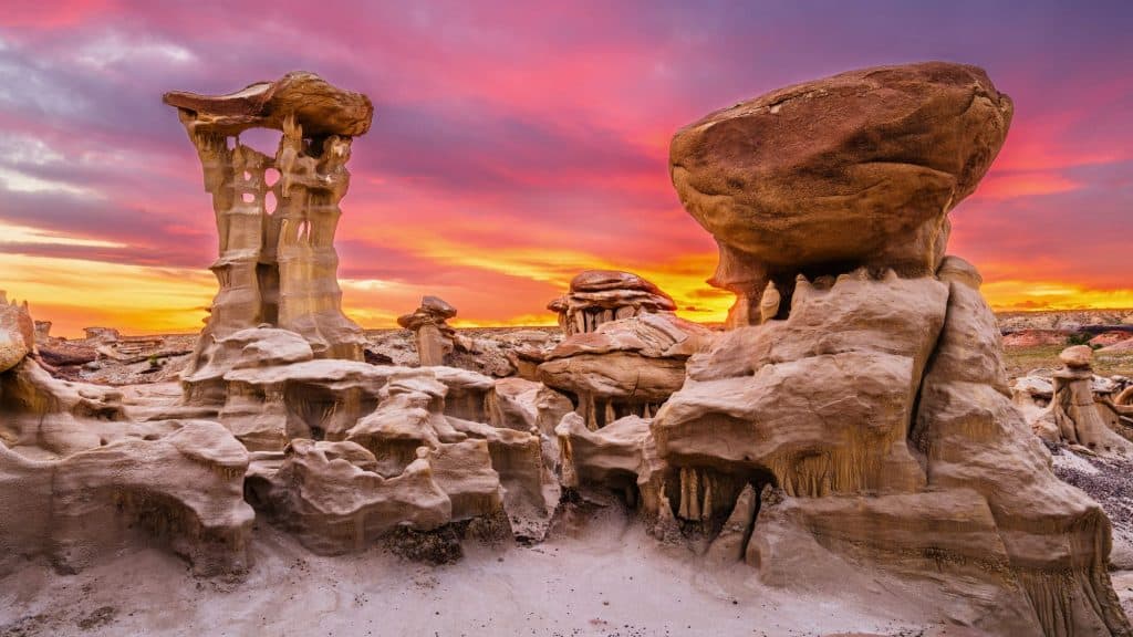 A surreal desert landscape in the Bisti De-Na-Zin Wilderness glows beneath a vibrant sunset, featuring intricately eroded hoodoos and strange, sculptural rock spires. The colorful sky adds a dreamlike backdrop to this already alien-looking terrain.