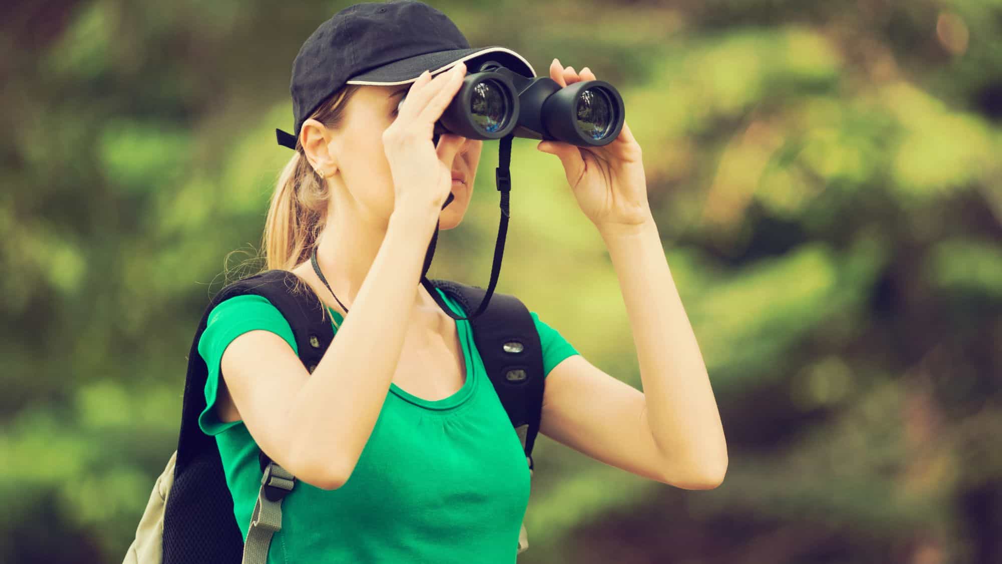 A woman wearing a green shirt, black cap, and backpack peers through binoculars in a lush forest, likely birdwatching or wildlife spotting during a hike.