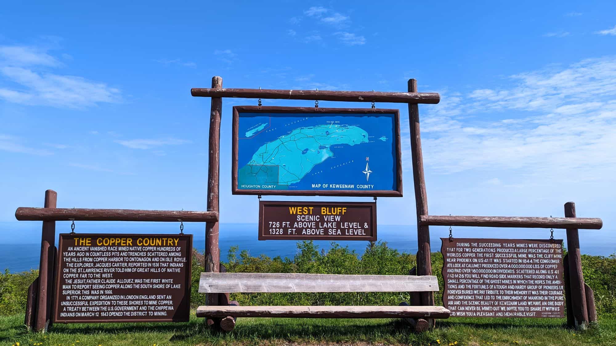 Wooden sign installation at Brockway Mountain displays a map of Keweenaw County and interpretive panels titled “The Copper Country” and “West Bluff Scenic View.” The sign explains the area's mining history and elevation—726 feet above lake level, with Lake Superior and a clear blue sky in the background.