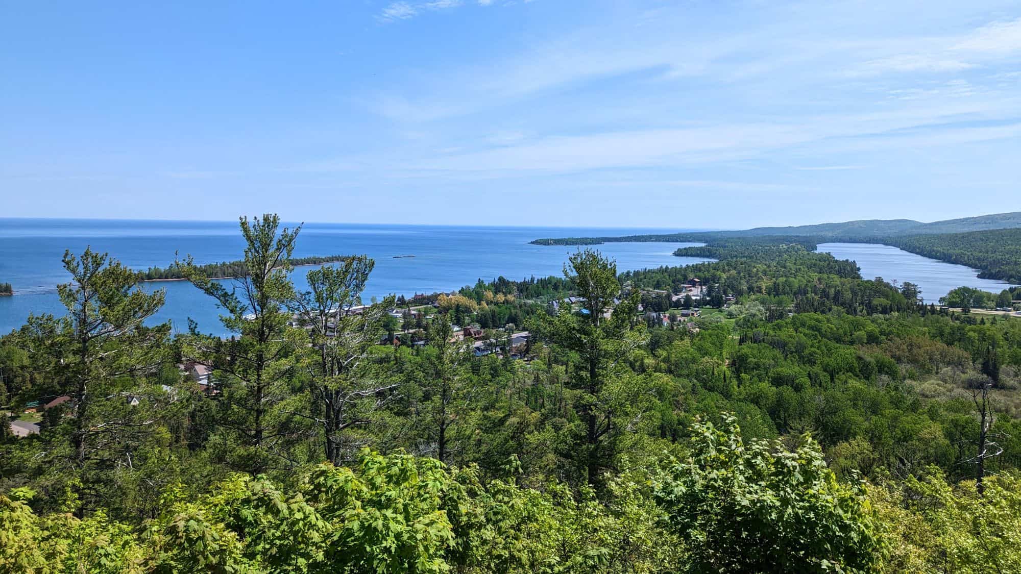 A scenic overlook from Brockway Mountain reveals a sweeping view of lush forest, a lakeside town, and Lake Superior stretching out to the horizon under a crisp blue sky.