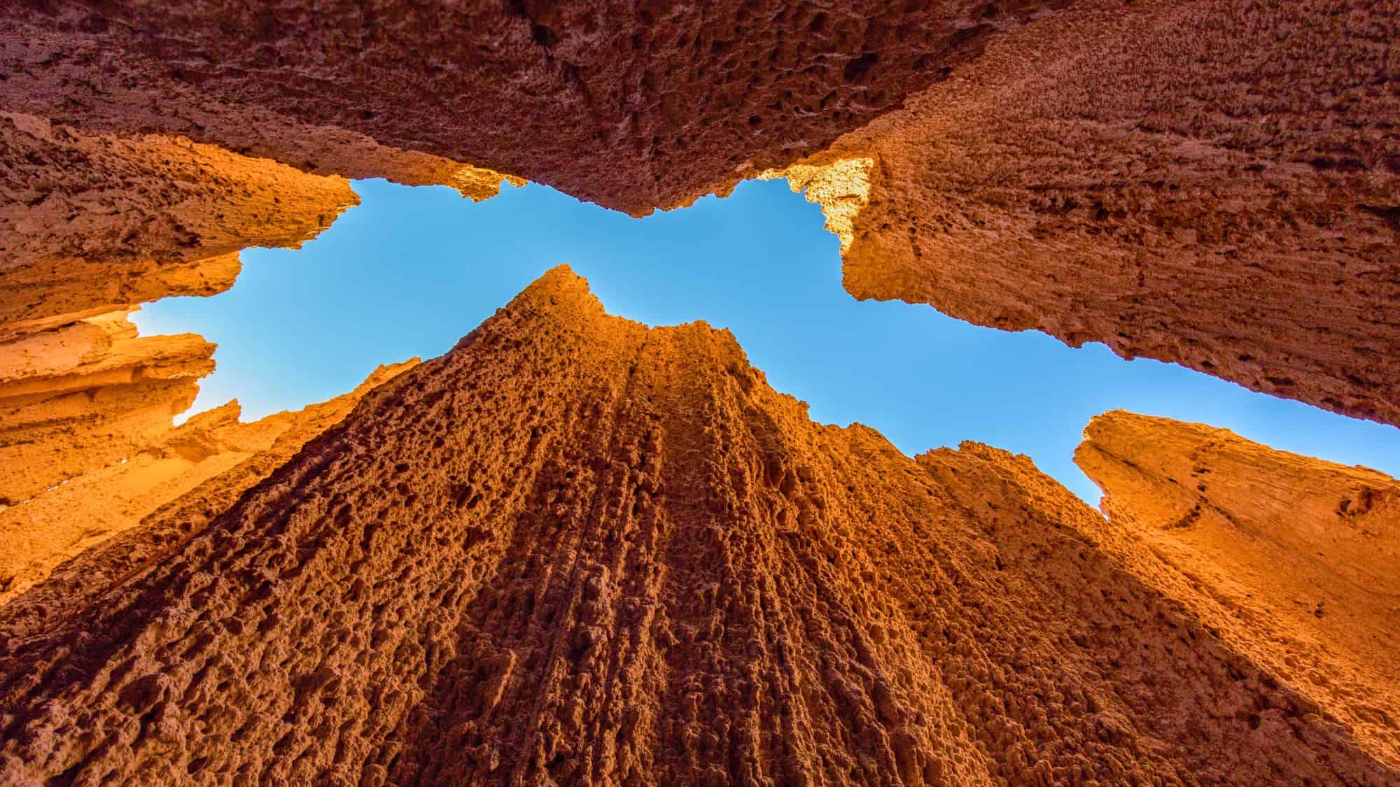 A dramatic upward view between tall, narrow red rock spires in Cathedral Gorge, Nevada, revealing a sharp blue sky above. The sun casts golden highlights on the rugged, textured rock walls.