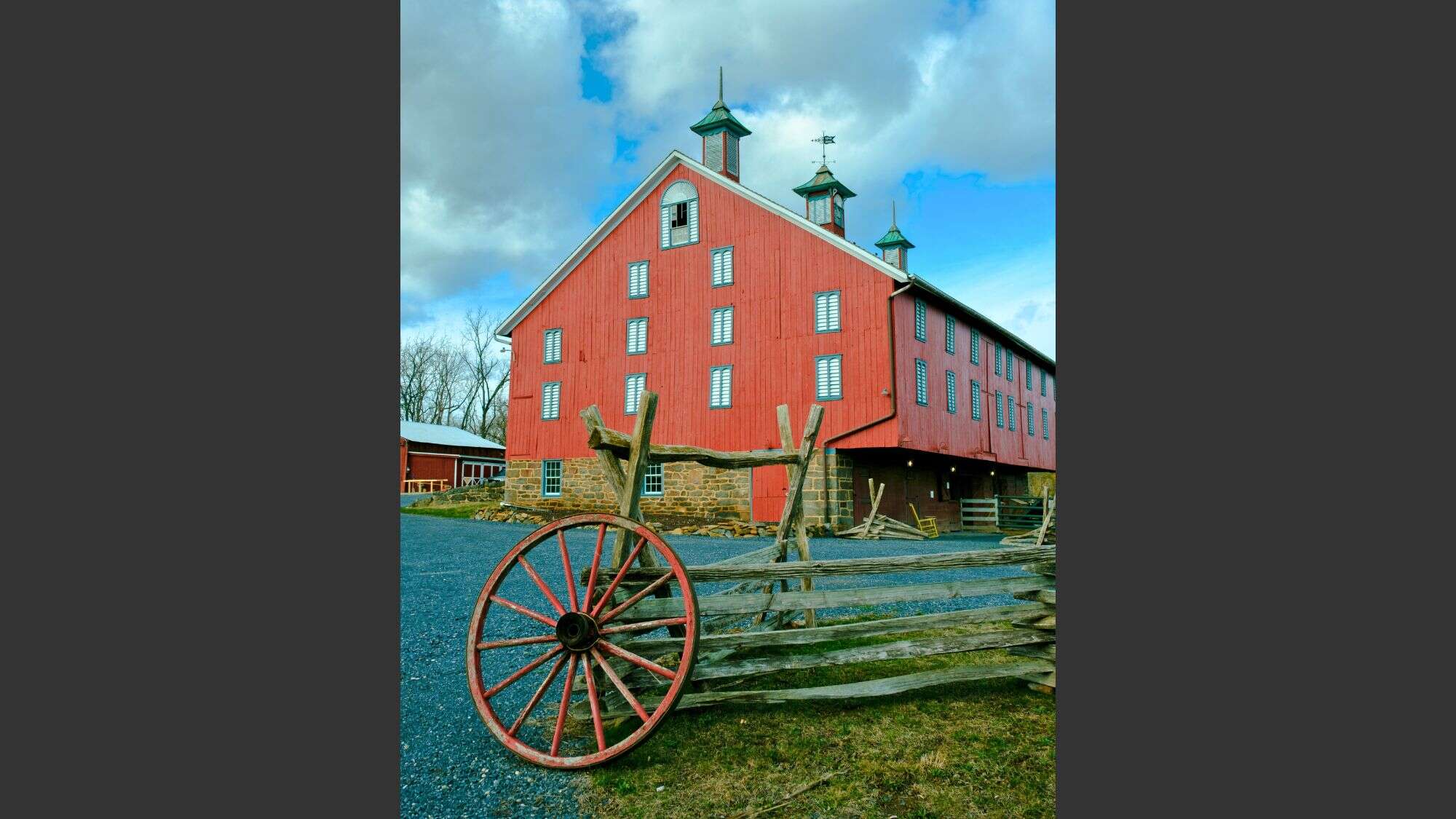 A historic red barn with stone foundation and multiple cupolas stands behind a rustic wooden fence and a red wagon wheel, under a partly cloudy sky.