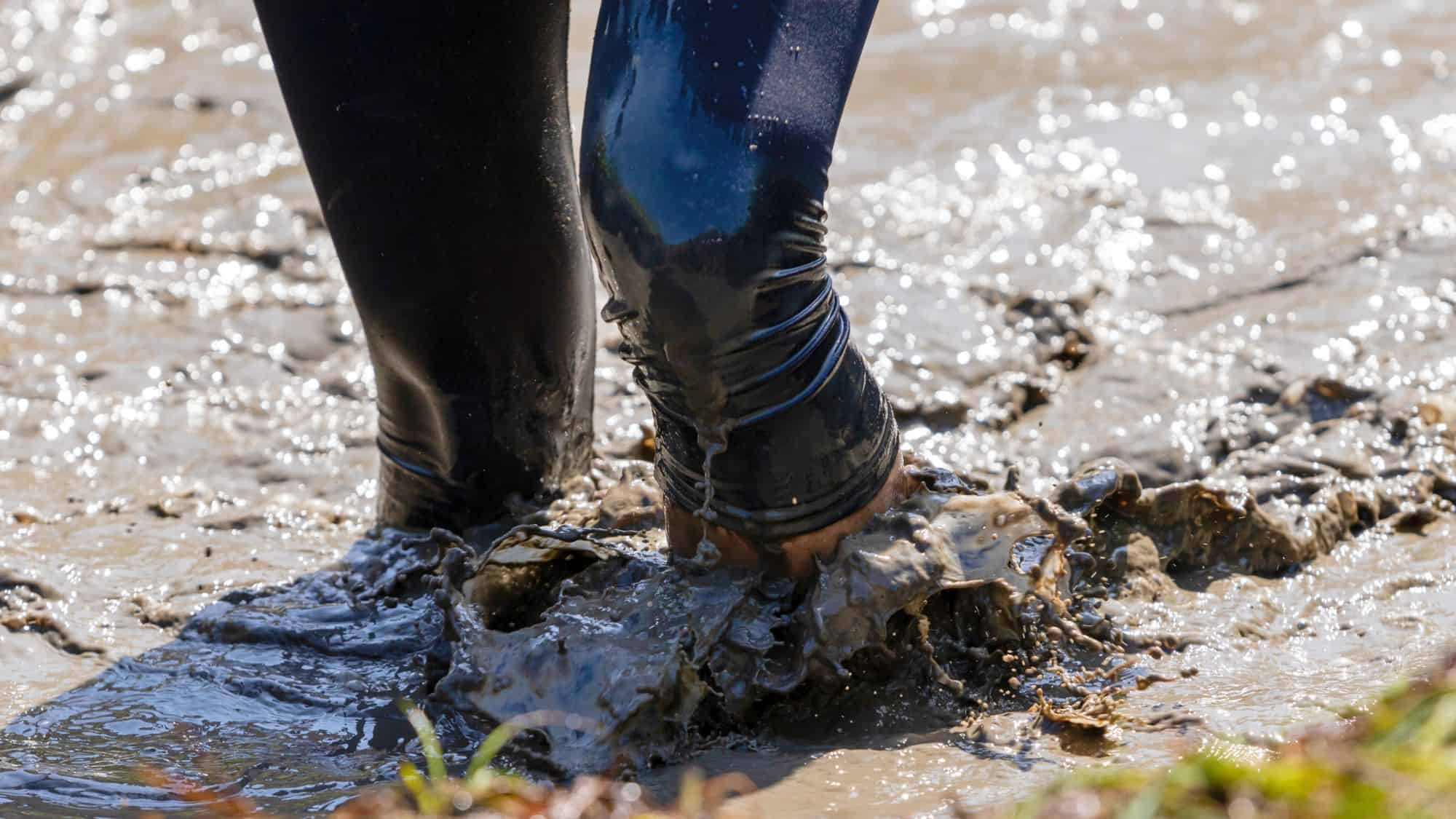 Close-up of a person’s legs stepping through deep, wet mud, with water splashing around as their feet sink, showing the messiness of a muddy trail.