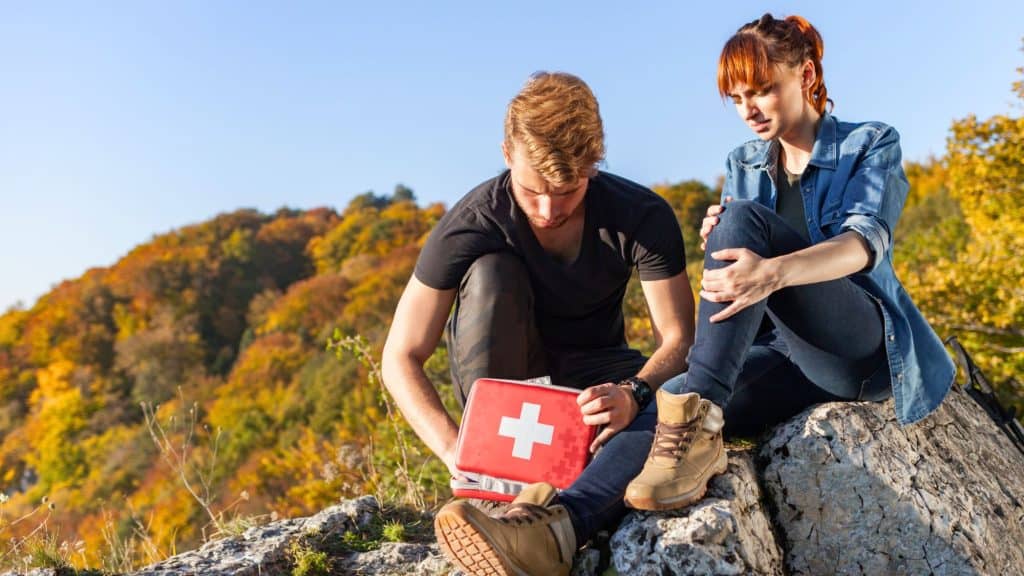 A man kneels beside a woman sitting on a rocky outcrop in the mountains, opening a red first aid kit as she holds her injured knee, surrounded by vibrant autumn foliage under clear blue skies.
