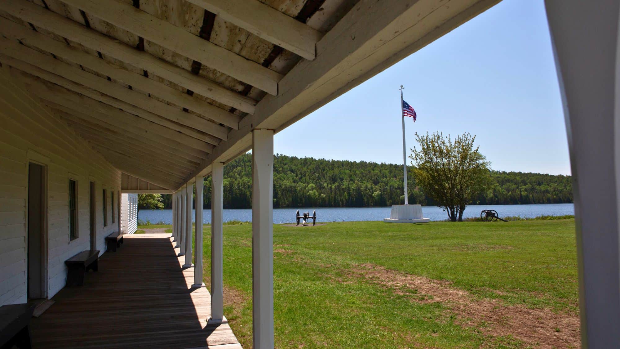 A view from the shaded porch of a historic white wooden fort building overlooking a grassy field, a flagpole with an American flag, a cannon, and a tree by the edge of a calm lake with forested hills in the background.