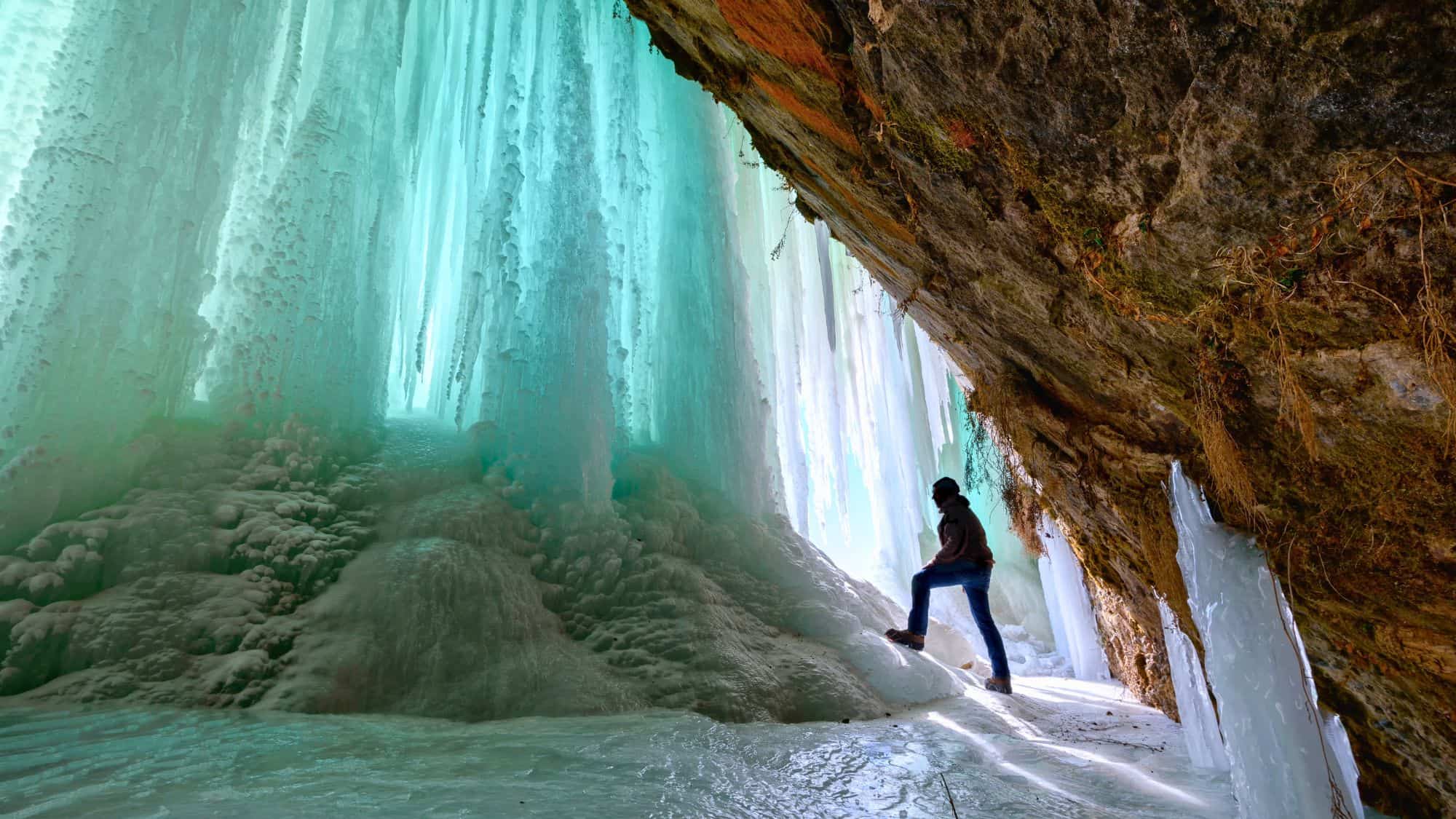 A hiker steps into a surreal ice cave near Munising, Michigan, surrounded by towering curtains of frozen turquoise icicles glowing with light filtering through the translucent ice.