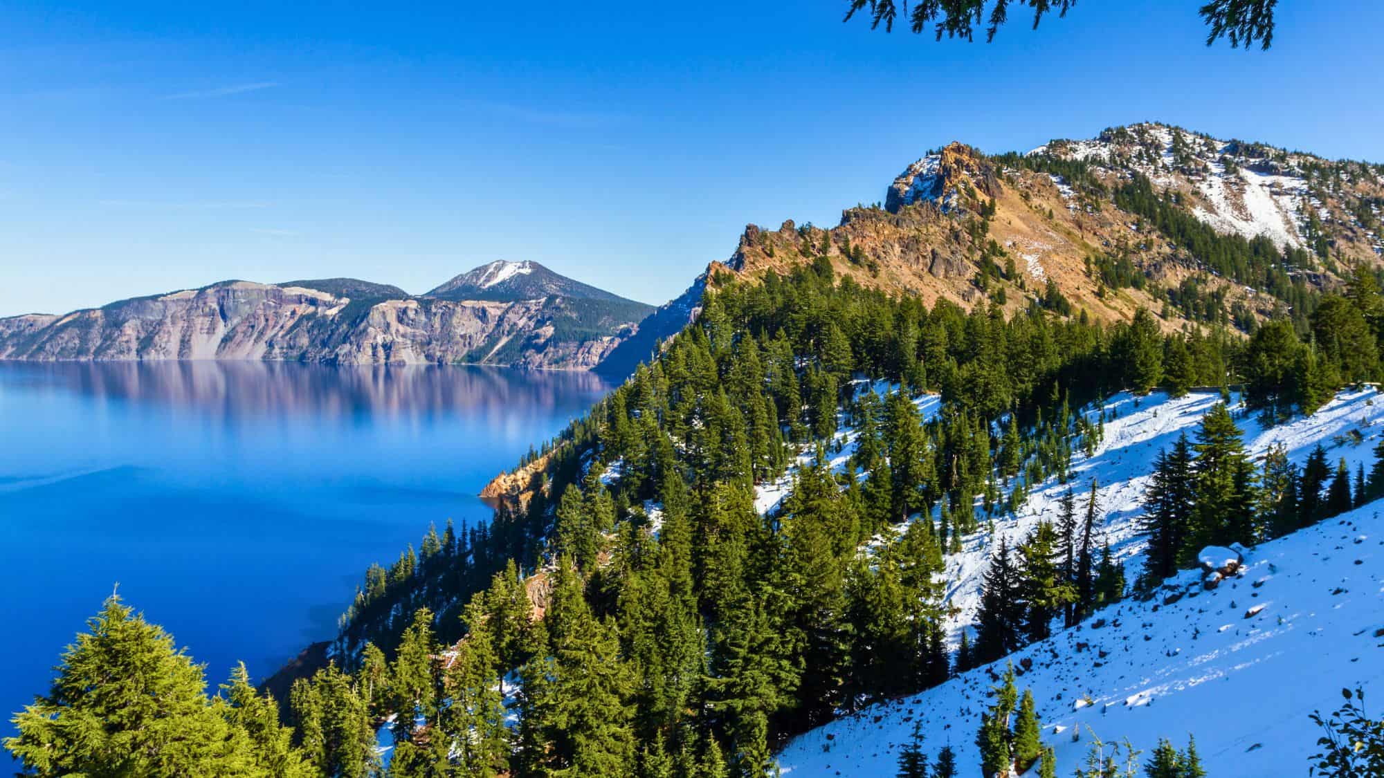 Panoramic daytime view from Garfield Peak overlooking Crater Lake, with evergreen trees, patches of snow, and the lake’s deep blue water reflecting the surrounding cliffs.