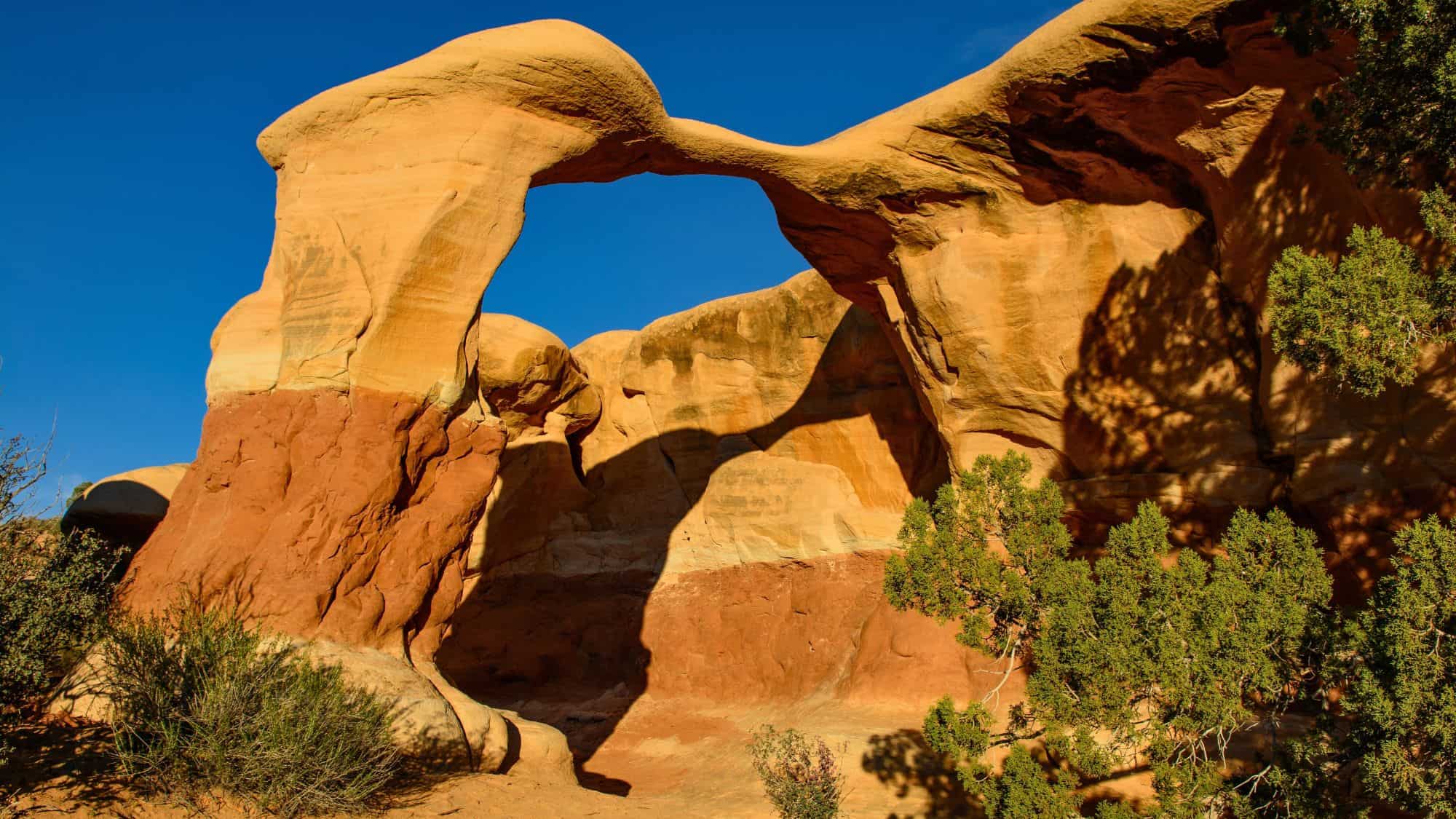 A natural sandstone arch stands against a deep blue sky in Utah, with soft red and tan hues in the rock and desert vegetation at its base. The arch casts a bold shadow on the canyon floor.