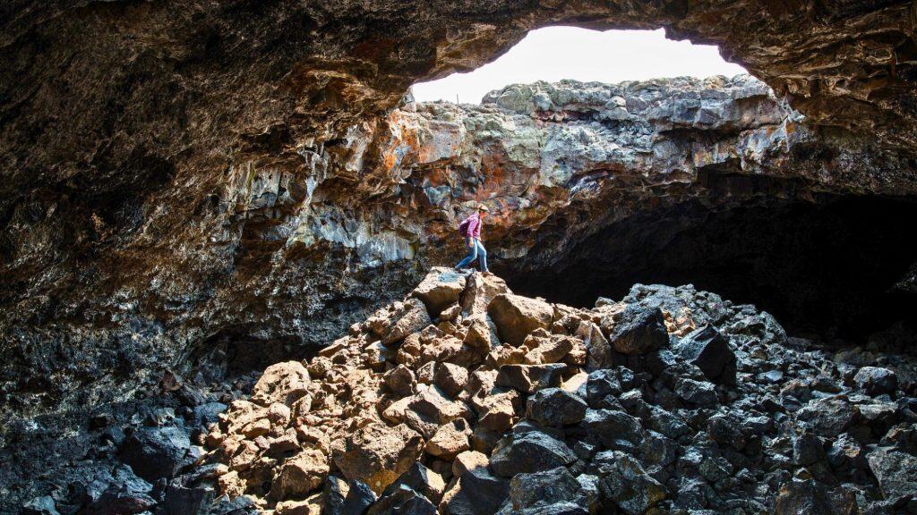 A person stands atop a pile of broken lava rock inside the Indian Tunnel, dramatically lit beneath a massive skylight that opens to the surface above.