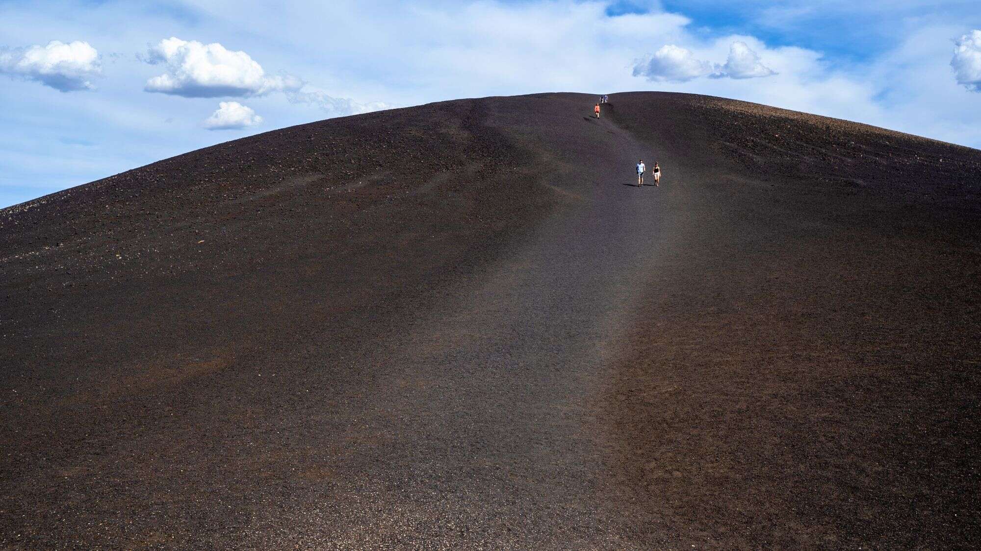 Several hikers trek up and down the steep, dark slope of Inferno Cone, a cinder cone volcano covered in black volcanic ash, under a blue sky scattered with clouds.