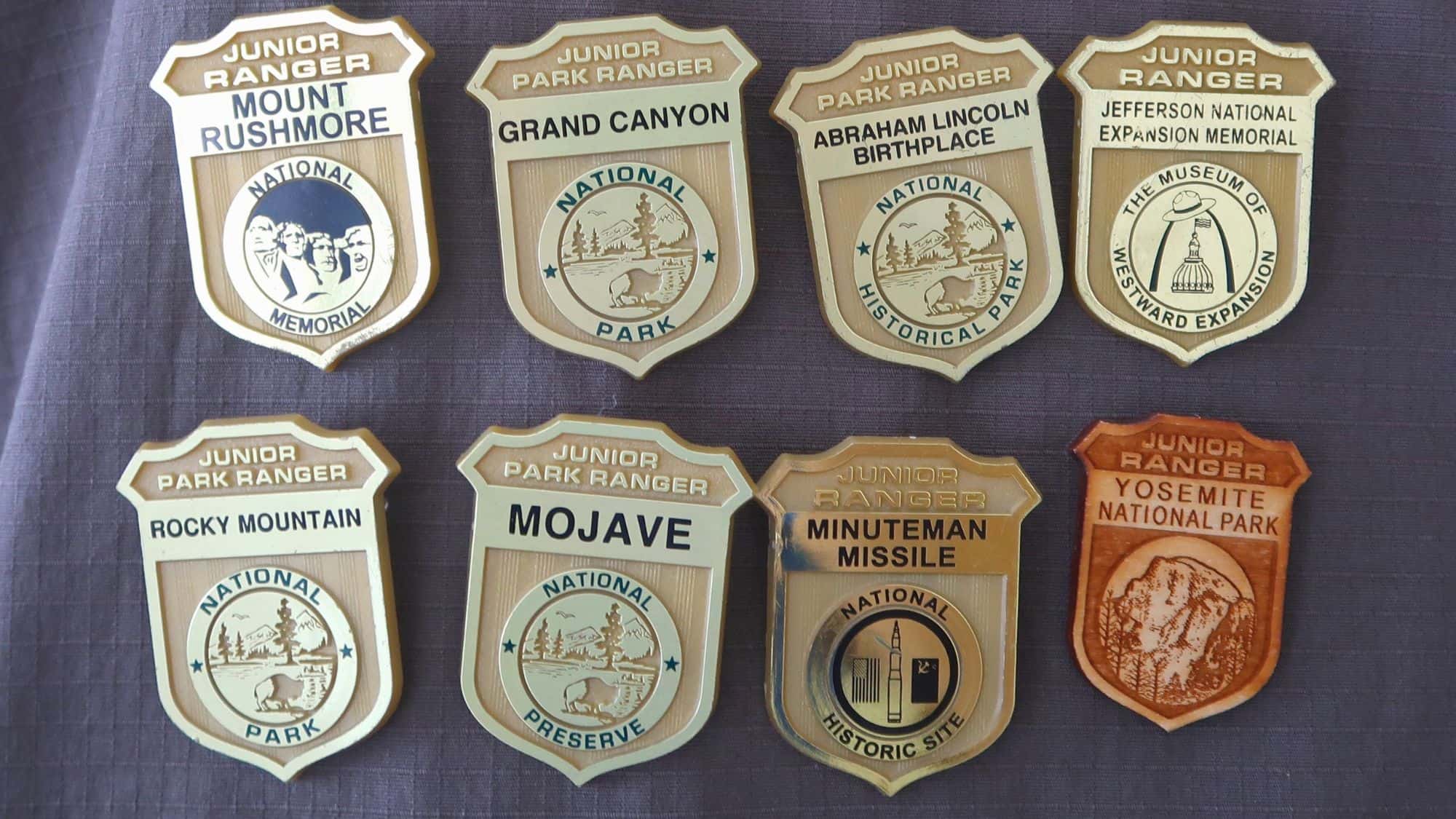 A collection of eight Junior Ranger badges from different U.S. national parks, including Mount Rushmore, Grand Canyon, and Yosemite, displayed on a gray fabric surface. Each badge has unique text and graphics reflecting its park.