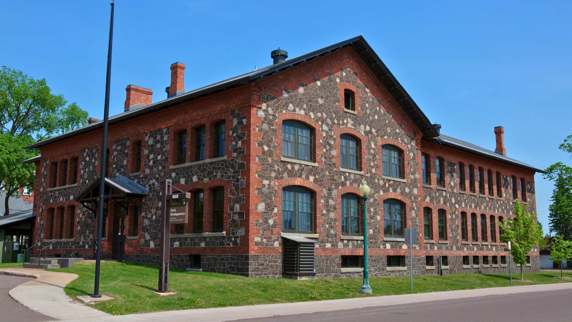 A large brick and stone building with arched windows houses the Keweenaw National Historical Park headquarters; the structure features historic architecture and sits along a quiet road under a bright blue sky.