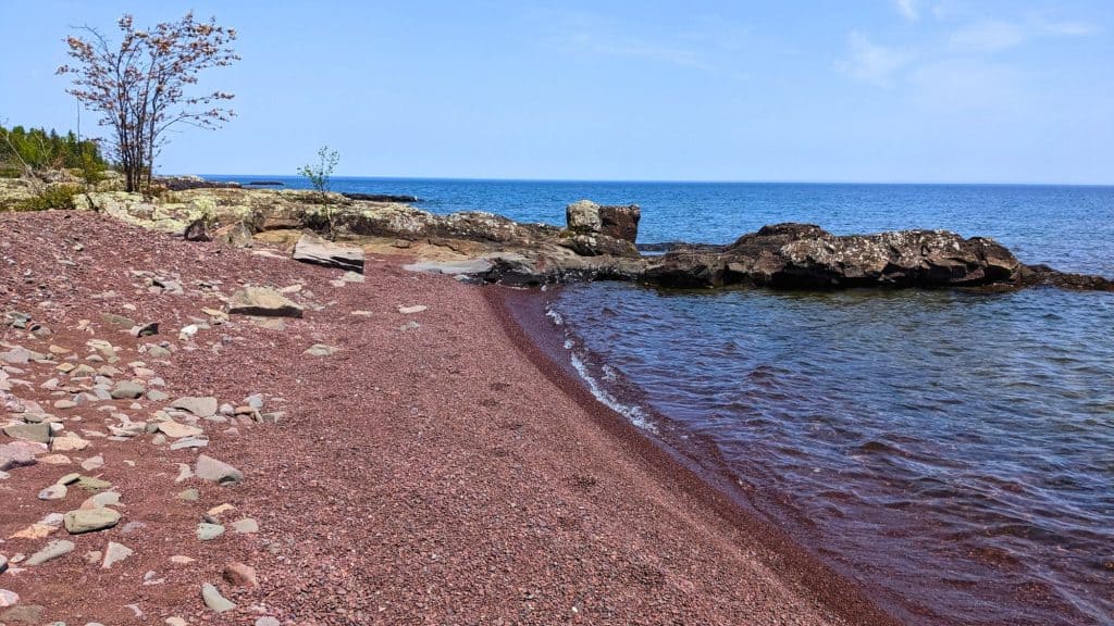 A rocky shoreline featuring a beach with reddish gravel meets the deep blue water of Lake Superior under a bright sky. Scattered stones and rocky outcrops add to the rugged coastal charm.