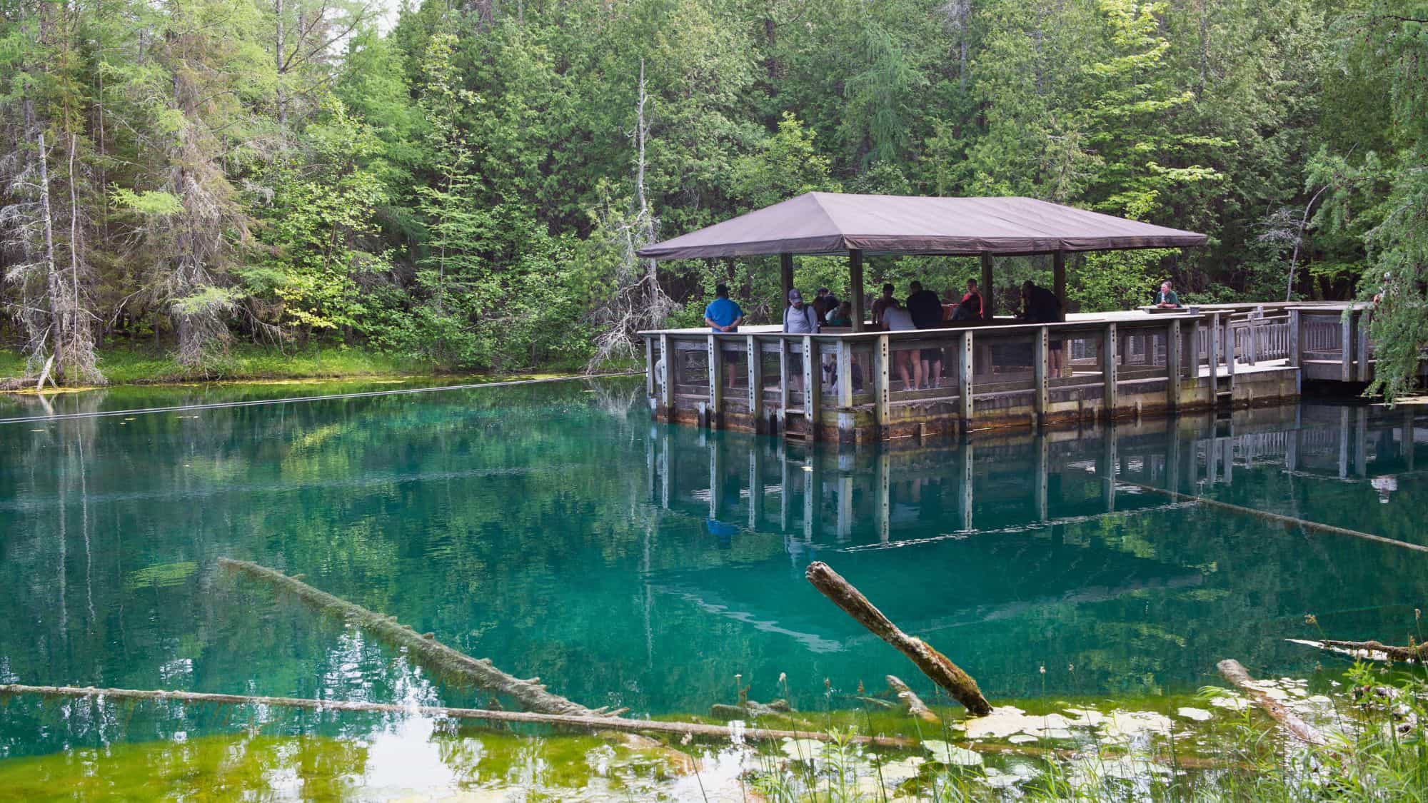 Visitors stand on a covered raft platform floating on the clear, turquoise waters of Kitch-iti-kipi, Michigan’s largest natural freshwater spring, surrounded by lush green forest.