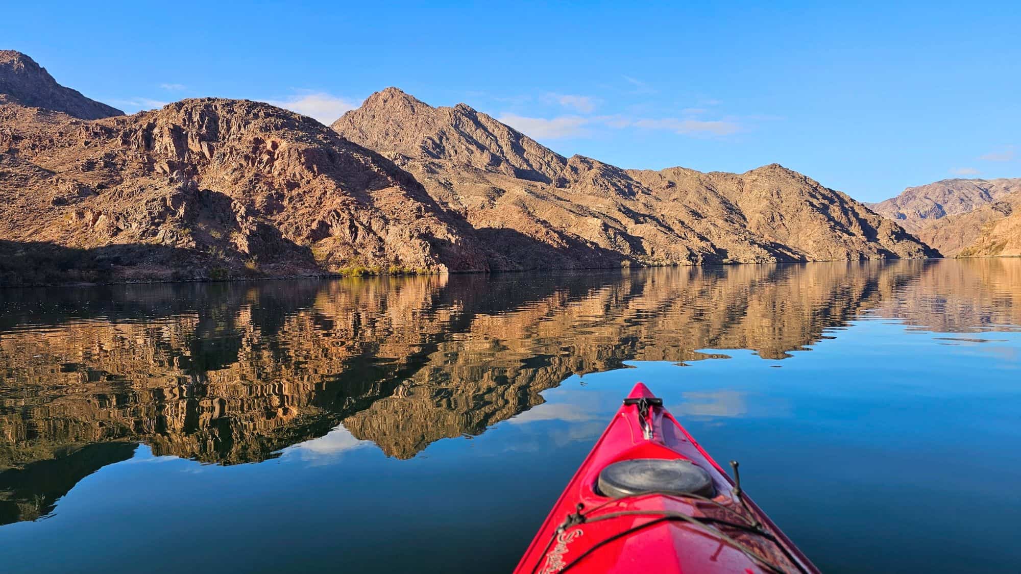 The tip of a red kayak points towards rugged, rocky mountains reflected perfectly in the still waters of Lake Mohave, under a vibrant blue sky.