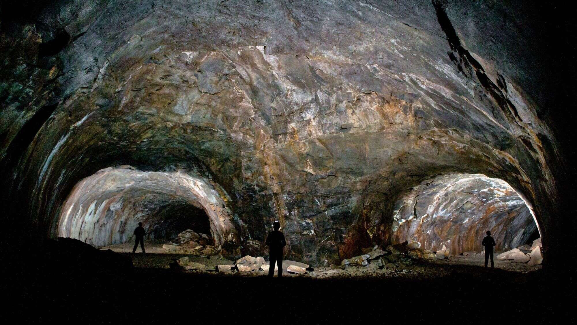 A wide-angle view of the inside of a dark lava tube cave with three silhouetted figures standing near its massive arched entrances. The rough rock walls are faintly lit by headlamps.