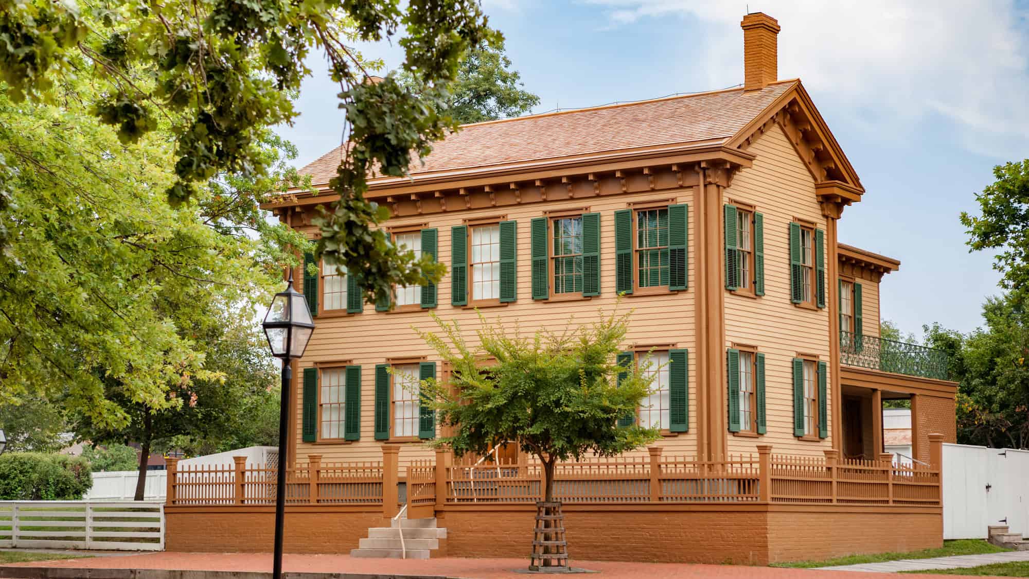 A well-preserved, two-story wooden house with green shutters and a picket fence sits on a tree-lined brick street in Springfield, once home to Abraham Lincoln.