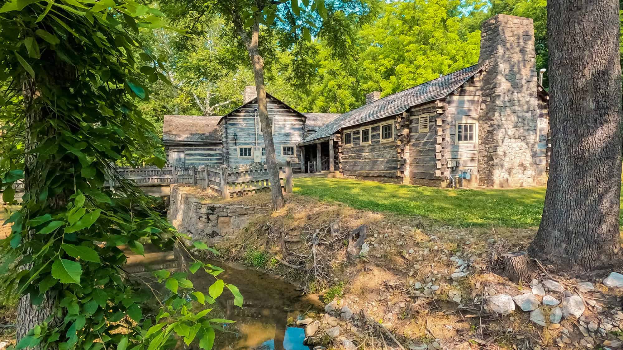 A rustic pioneer village cabin made of logs and stone chimney sits tucked in a forested area, bordered by a small creek and shaded by tall trees.
