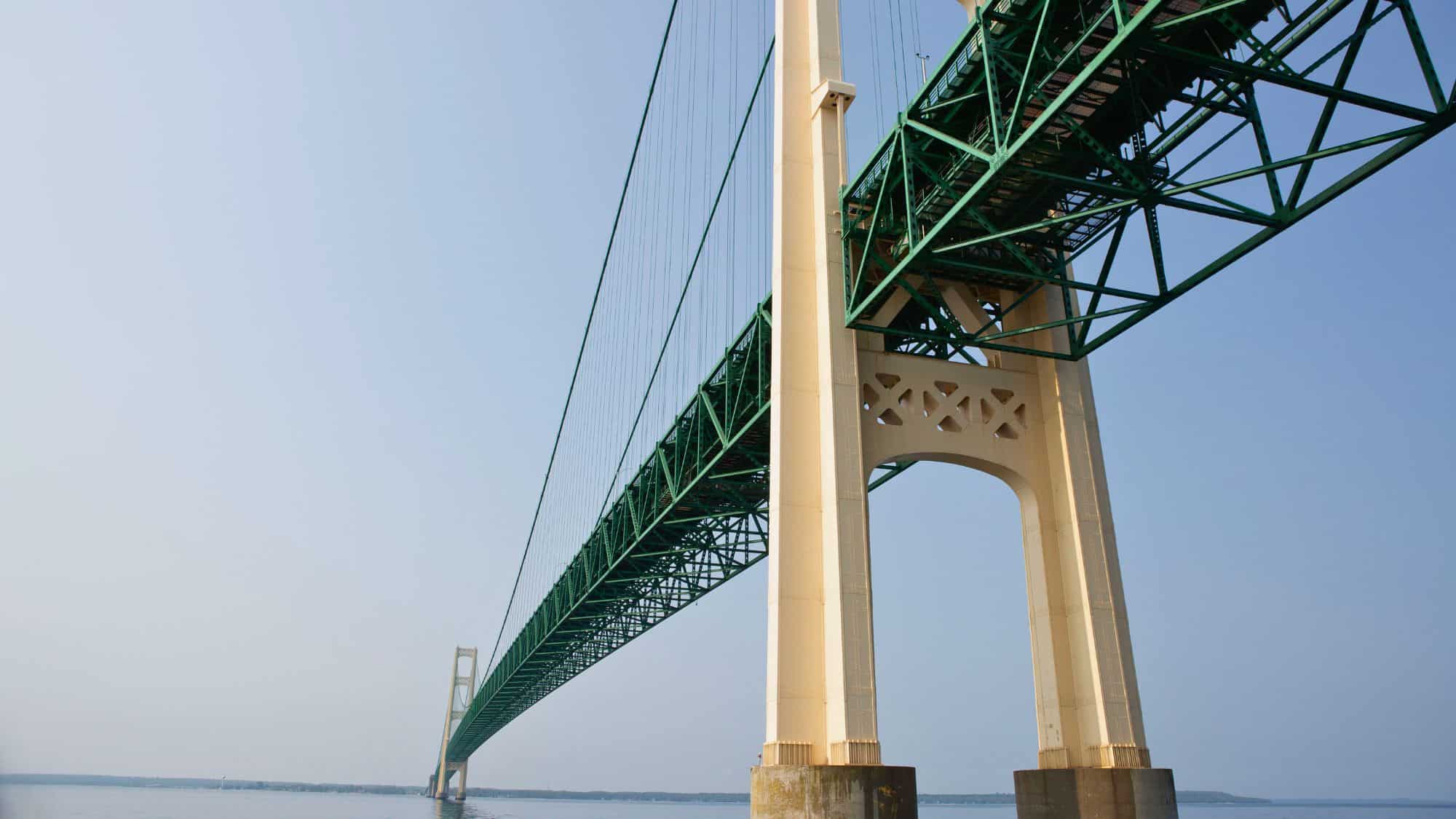 An upward perspective of the Mackinac Bridge, showcasing its tall concrete pillars and green steel framework stretching across the water. The image captures the bridge's impressive engineering from below.