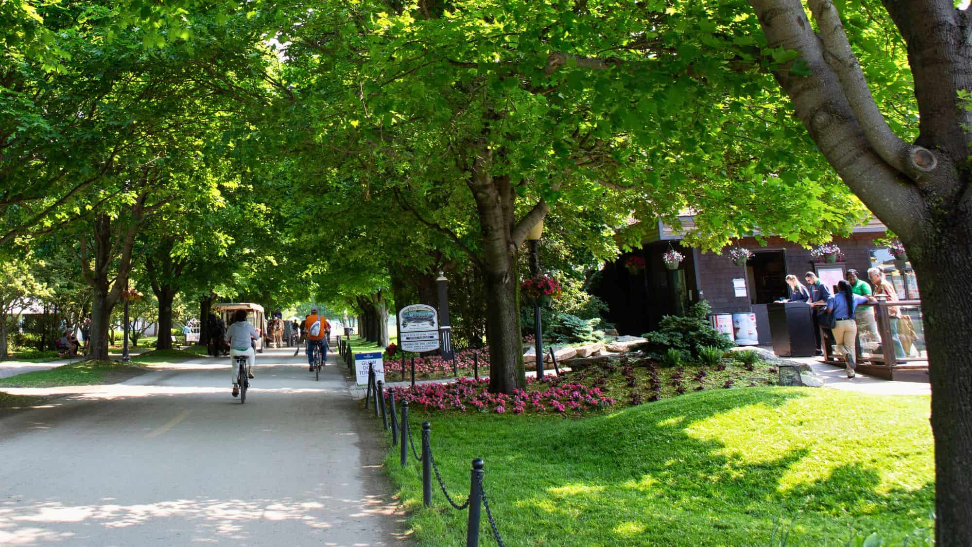 People bike and walk beneath a shady canopy of trees along a paved pathway on Mackinac Island, with flowerbeds, signs, and horse-drawn carriages adding charm to the lively scene.
