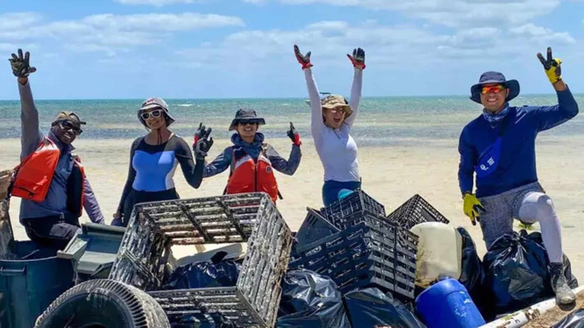 A group of smiling volunteers pose on a sandy beach with piles of collected trash, including plastic bins, bags, and tires. They flash peace signs and cheer under a sunny sky near turquoise waters.
