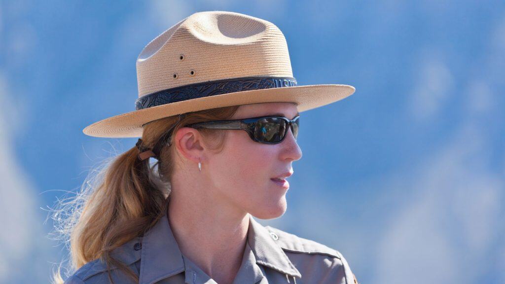 A female park ranger wearing a wide-brimmed hat and sunglasses looks to the side against a soft blue sky, with her park uniform crisp and official.