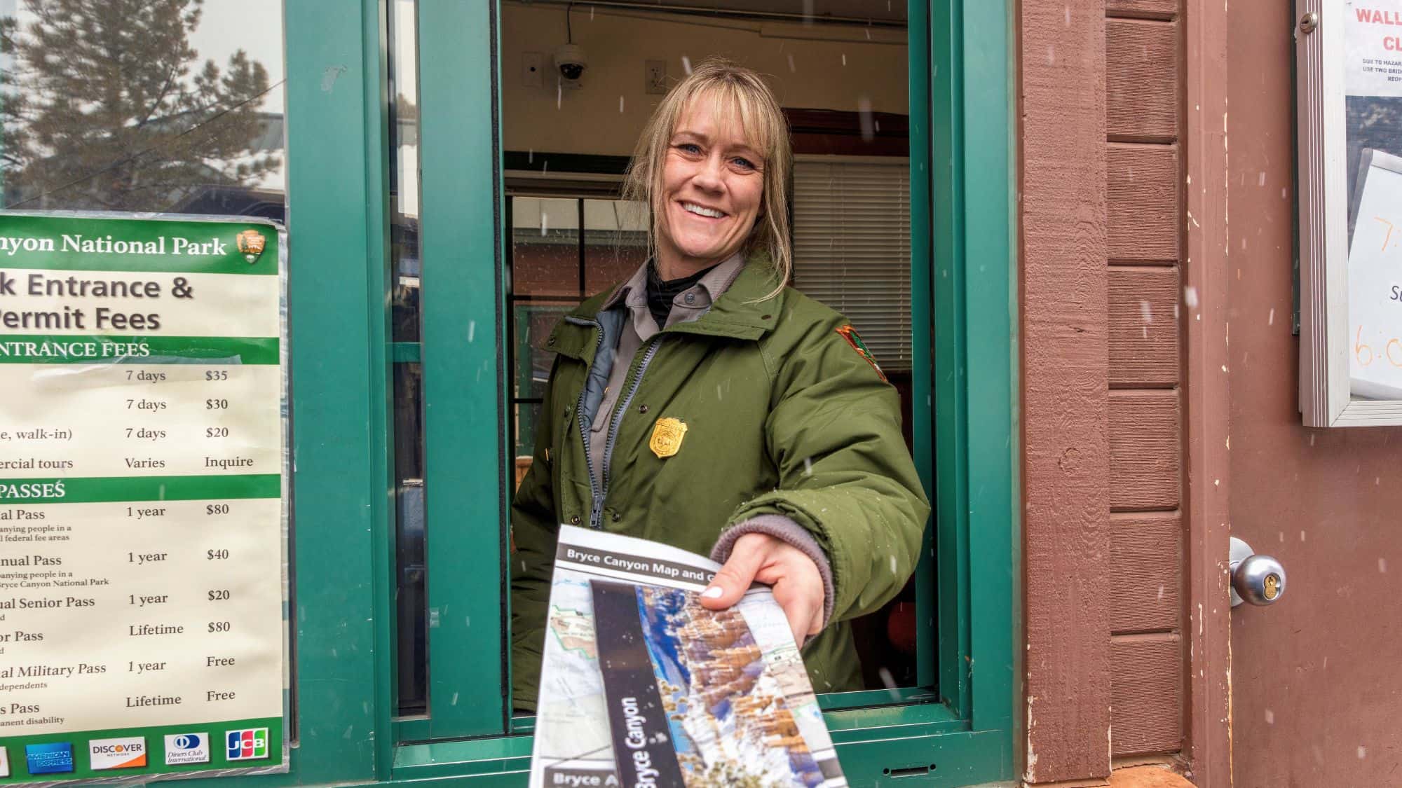 A friendly park ranger in a green uniform and jacket hands out a Bryce Canyon National Park map from a booth window. A price list for entrance and permit fees is visible on the booth’s exterior.