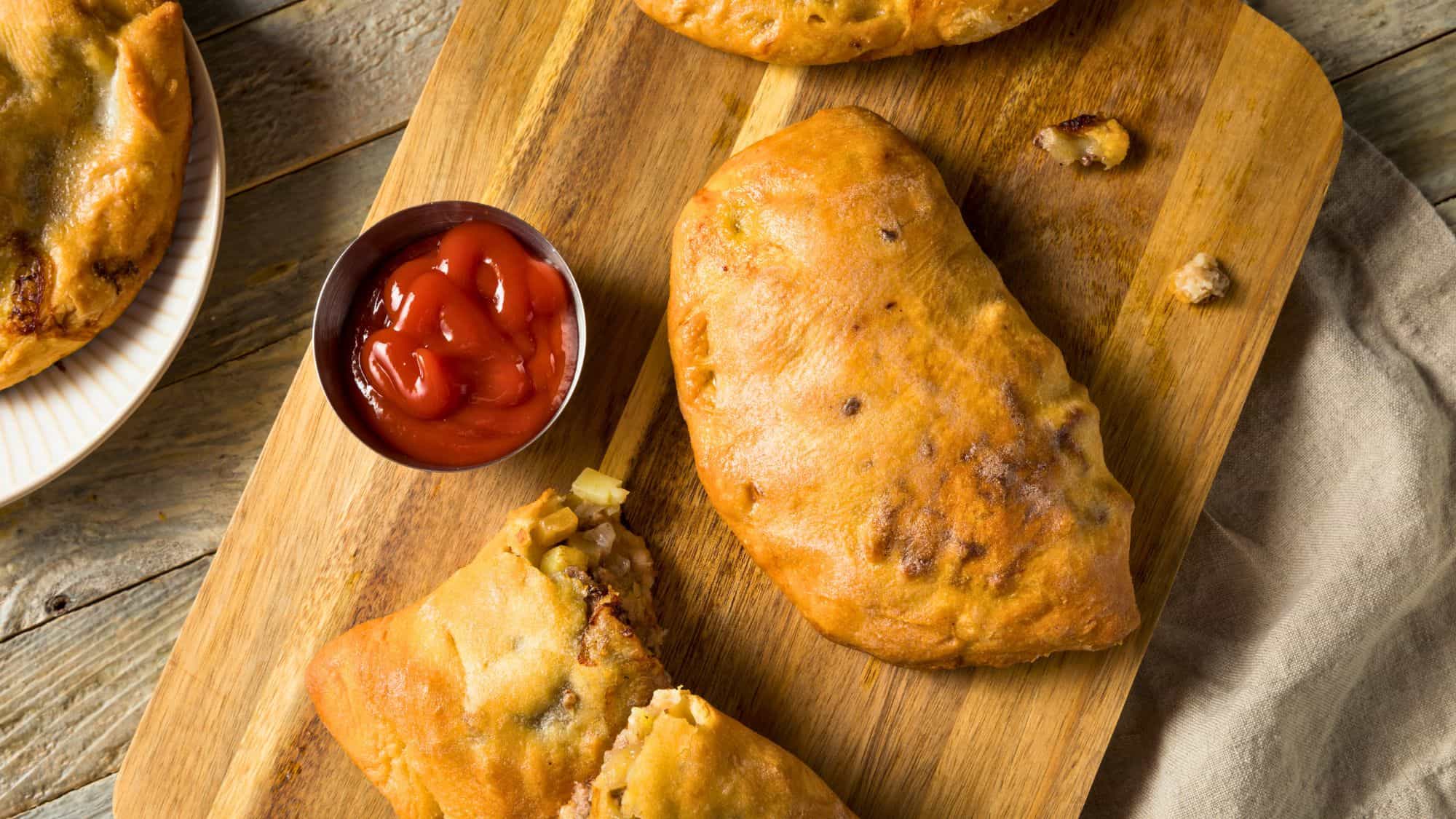 A golden-brown pasty sits on a wooden board, one of them cut open to reveal a hearty filling of meat and potatoes. A metal cup of ketchup sits nearby for dipping.