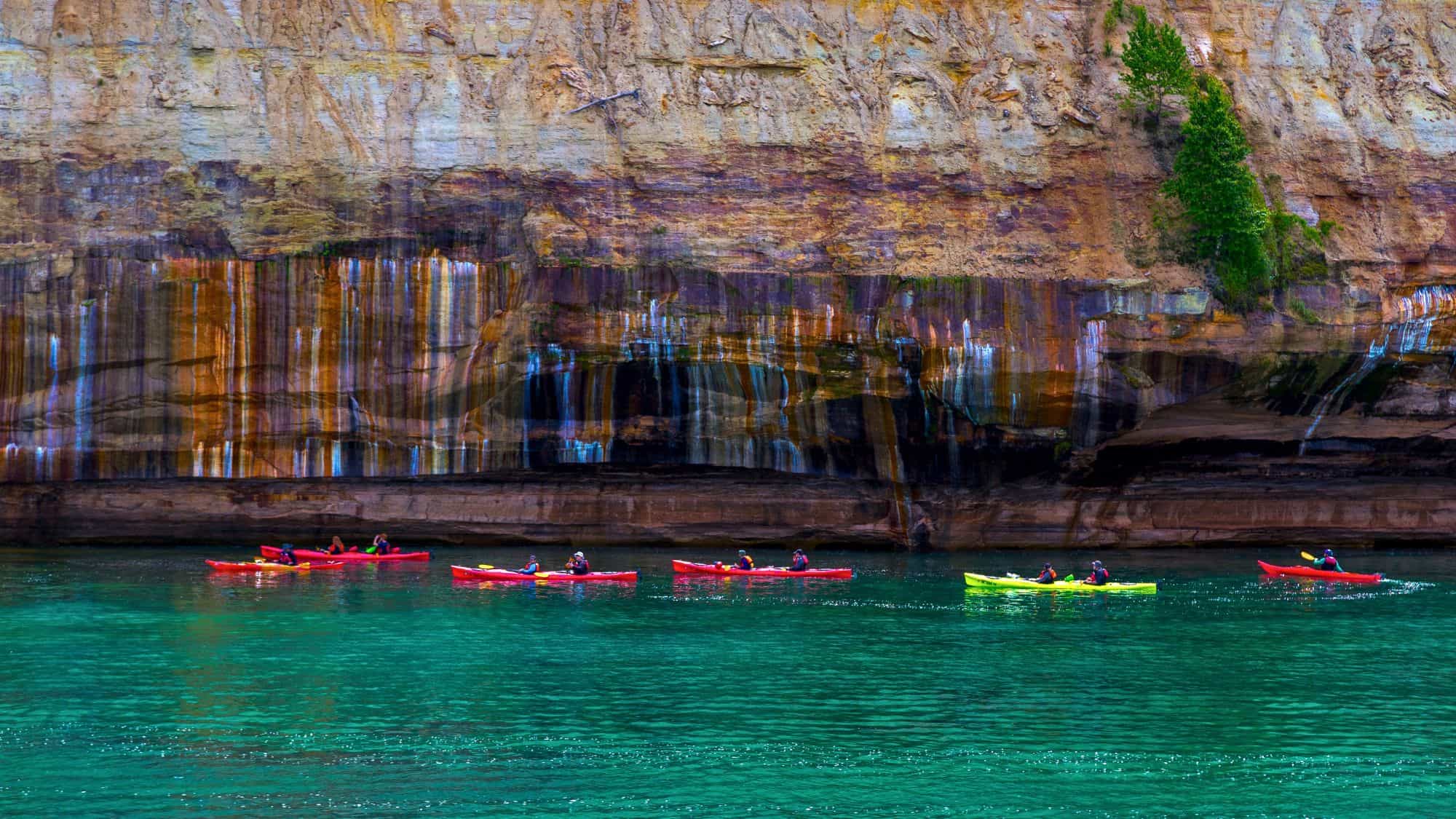 A line of colorful kayaks glides through turquoise water beside the striped mineral-stained cliffs of Pictured Rocks National Lakeshore. The contrast between water and rock makes for a striking natural scene.