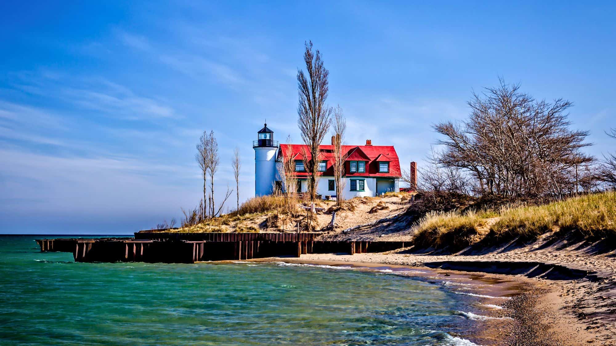 A striking white lighthouse with a cylindrical tower and a red-roofed keeper’s house sits atop a sandy dune along Lake Michigan. Surrounded by sparse trees and framed by turquoise waters and blue skies, the historic Point Betsie Lighthouse offers a picturesque coastal view.