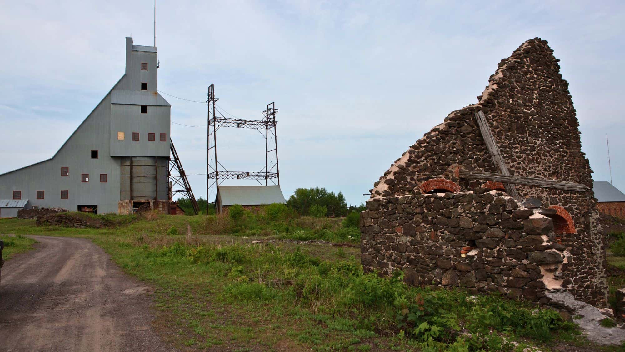 The historic Quincy Mine complex features a tall gray hoist house structure and ruins of stone buildings surrounded by grassy terrain and power lines under an overcast sky.